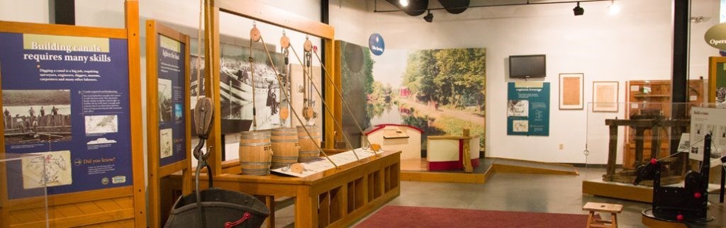 Interior of museum gallery with various artifacts and displays including pulley systems. Museum text panel at left reads "Building canals requires many skills"