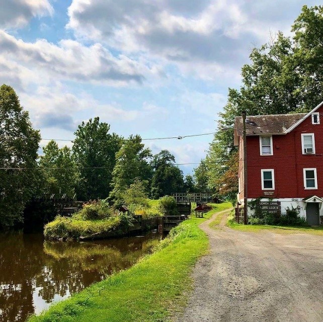 Red house with white trim at right. In center, gravel road. At left, a canal. Greenery all around. Blue sky with bright clouds above.