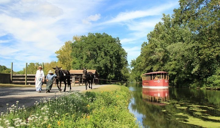 Two mule pull boat red canal boat down canal, accompanied by two people in historic costumes. Green trees on both sides of canal, and small white flowers in foreground. Sunny skies above.