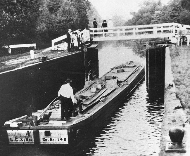 Historic black-and-white photo of long, flat barge passing under white wooden bridge on canal. One man stands on barge, several watch above on bridge and along canal locks. Grass and trees along canal