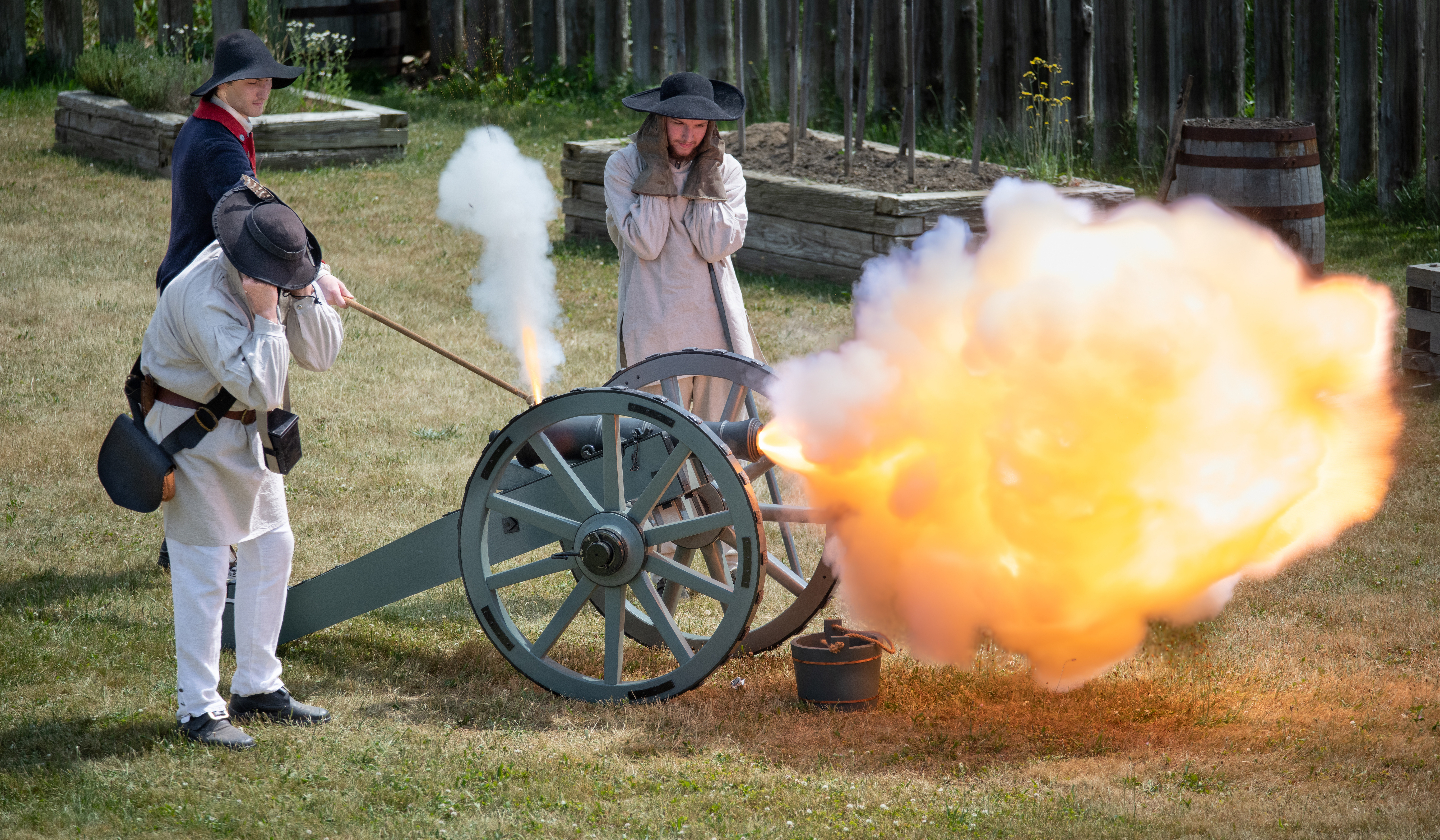 A cannon explodes a ball of fire while several people near it brace themselves from the shockwave.