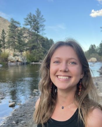 Blonde haired blue eyed girl smiling with a river, rocks, and trees in the background.