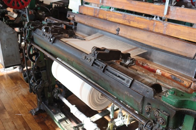 a roll of white cloth with a brown stripe on either side. The roll is situated on the underside of a metal machine, with the unrolled end feeding into the machine.