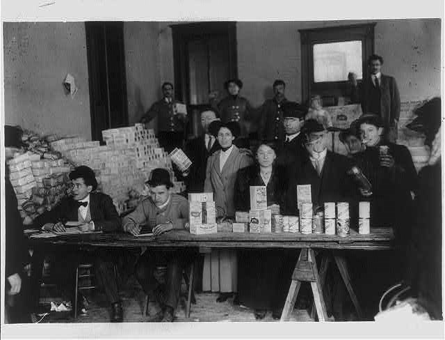 Several people sit and stand behind a table with boxes of food on it in a black and white photo.