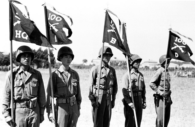 Five men in military uniforms holding flags for the 100th Infantry Battalion