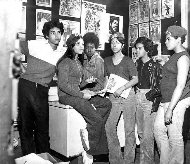 A Black and white photograph of six leaders of the Young Lords. They stand in a corner with art prints, posters and images. The figures are dressed in jeans and casual shirts. One member is wearing a beret and is holding papers