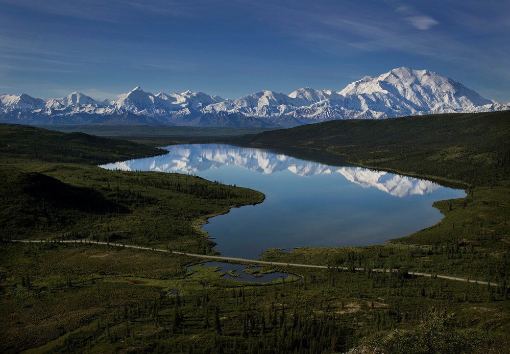 A large, calm lake reflecting glaciated mountains of the Alaska range in its water.