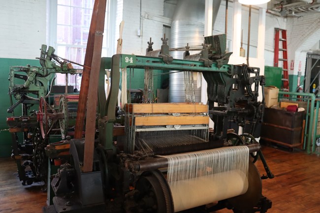A large metal machine painted green. A large beam holds threads that feed into the machine. Several wood and metal frames hang from a bar on the top and the threads feed through them. To the right there is a row of many metal arms in the upwards position