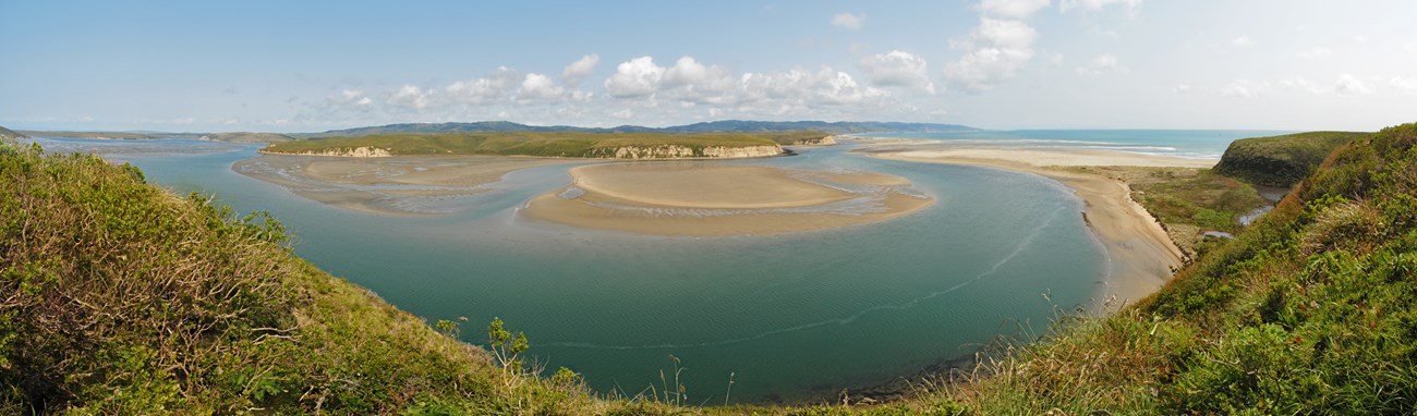 Panorama of channels of turquoise water between beaches and sandbars, connecting to the ocean. Light rocky bluffs and green hills surround the estuary.