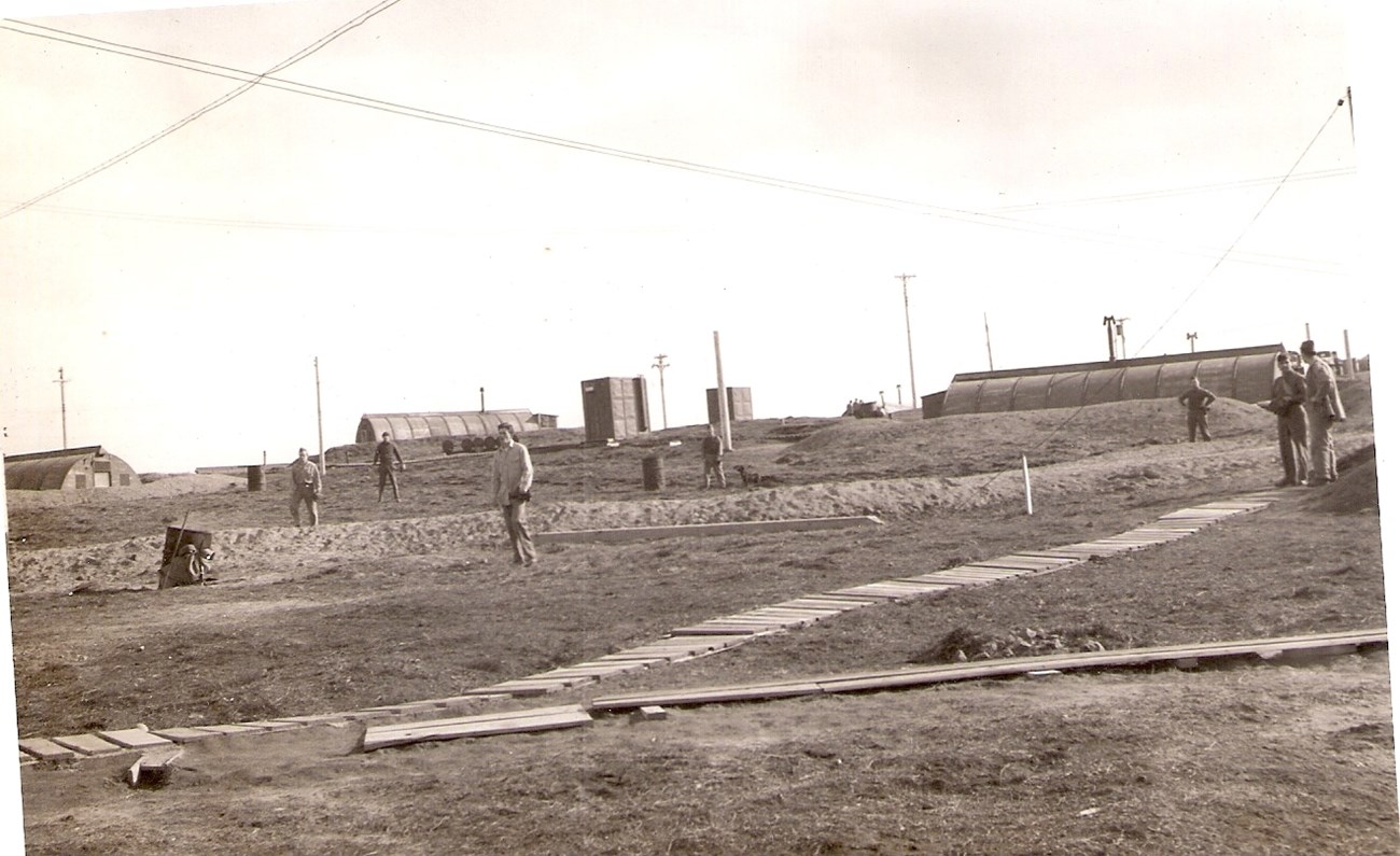 men scattered in grassy areas around Quonset huts and wooden boardwalks.
