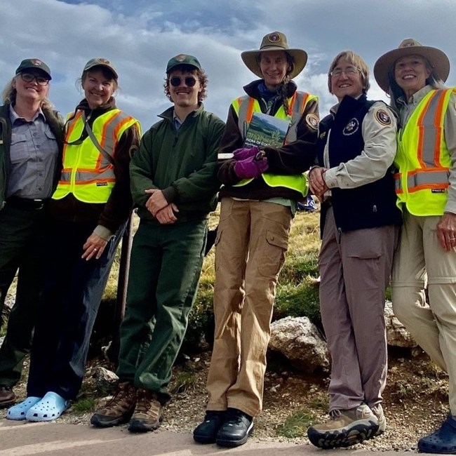 6 adults smile in front of natural landscape, two with ranger uniforms and four wearing volunteer hats and shirts.