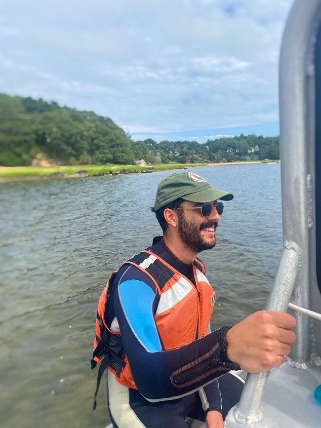 Scientist holds onto the edge of boat wearing wetsuit, hat, and sunglasses.