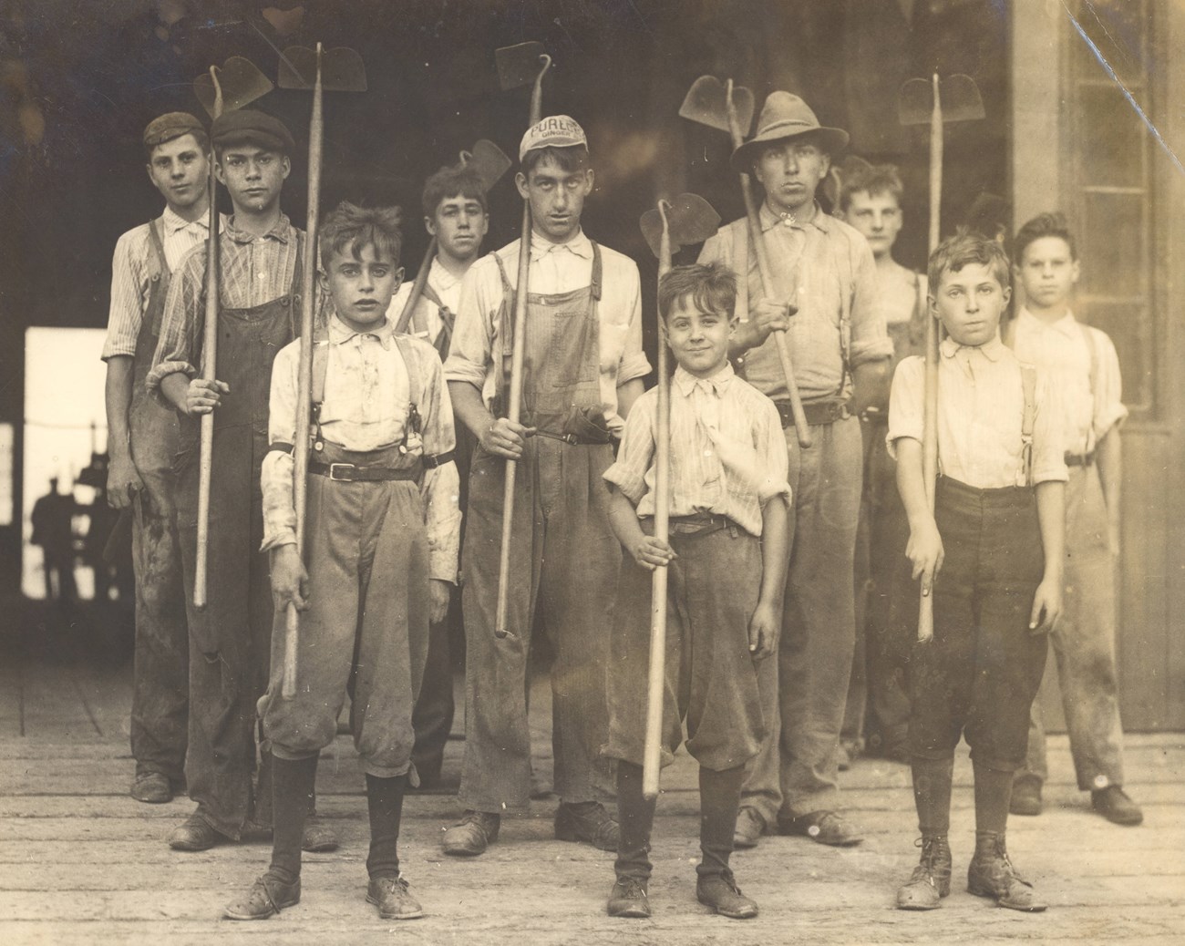 10 boys stand holding shovels and hoes resting on their right shoulders. They wear coveralls and buttoned and collared shirts.