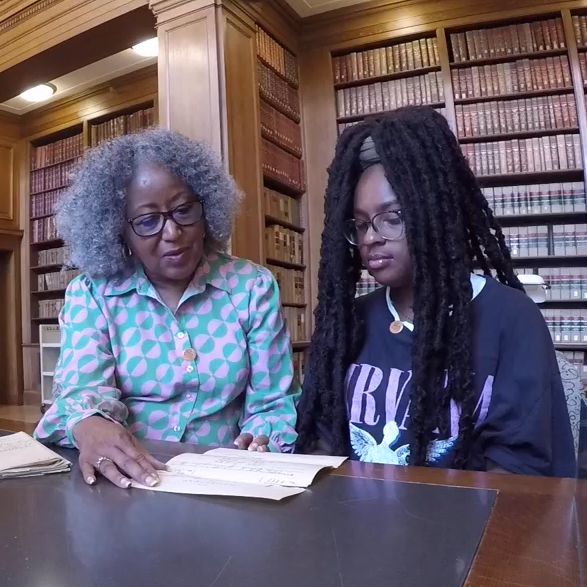 A woman shows a young adult a document in a library setting.