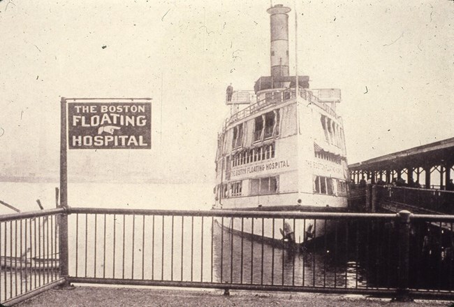 end of a pier with a sign Boston Floating Hospital and a multi-story steamer behind it in the water.