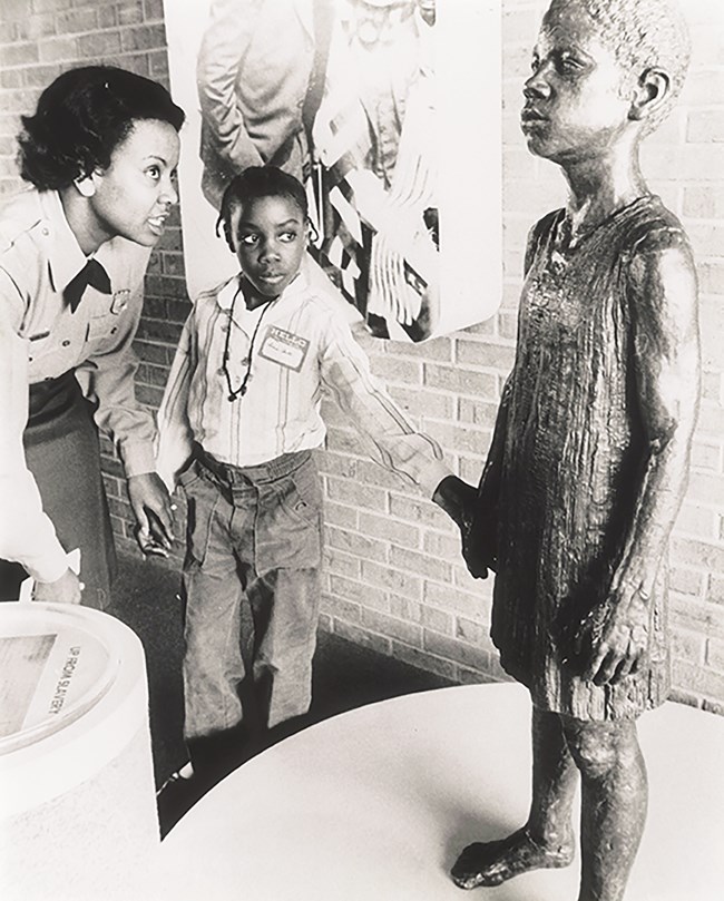 Geraldine Bell holds hands and bends towards a boy as he also holds the hand of a statue of Booker T. Washington as a boy.