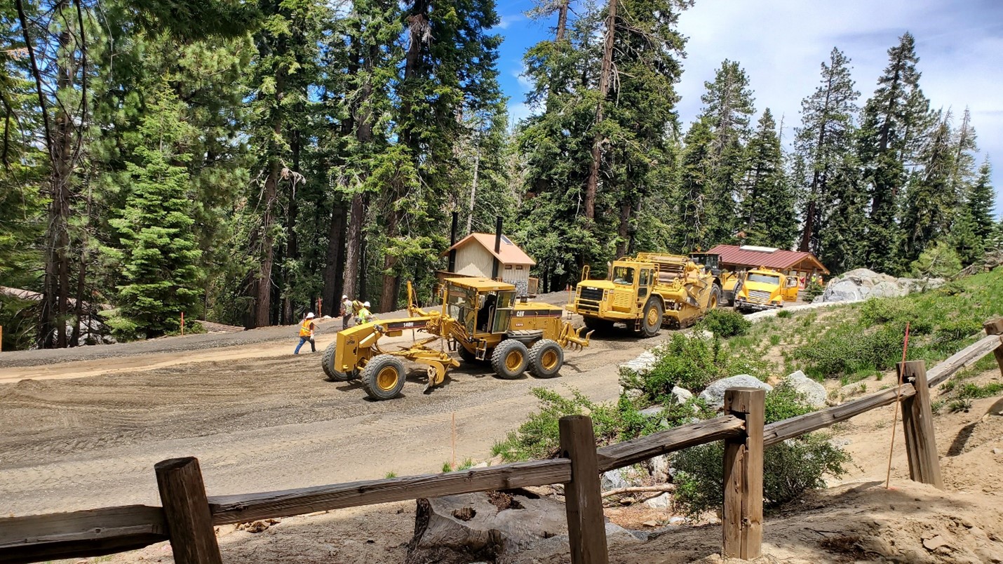yellow construction vehicles are parked along a dirt road. Tree and construction workers are in the background