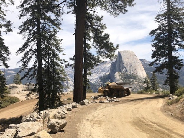 A yellow dump truck goes down a downhill curve on a mountainside. Blue skies and a few trees are in the background
