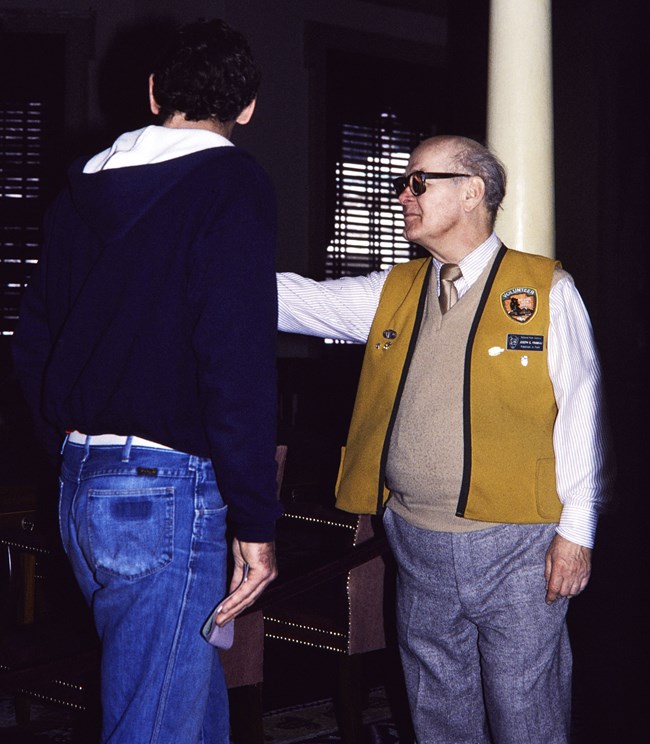Man in gold vest points in conversation with another man
