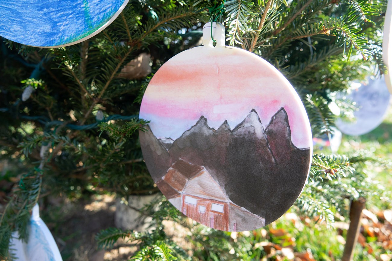 ornament with a drawing of mountains with snow and a house, portraying grand teton national park. The ornament is hanging from christrmas tree