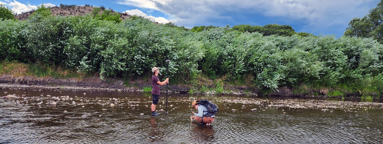 One scientist bent down to sample water and another scientist holding a water sampling equipment on a long pole attached to a tablet.