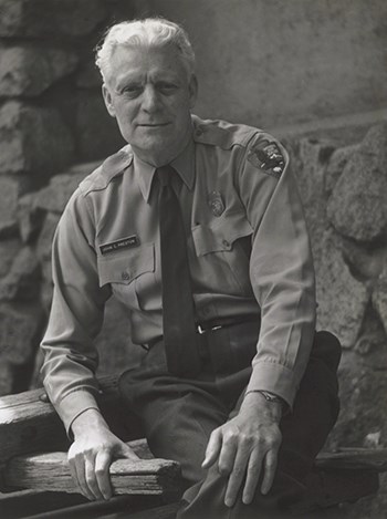 NPS Uniform Committee Chairman John C. Preston wears a long sleeved shirt, tie and name tag.  The NPS arrowhead patch can be seen on his left arm.