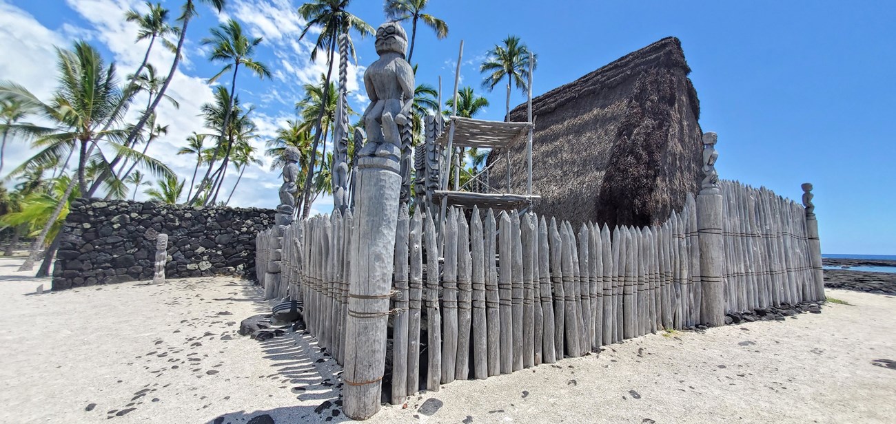Surrounded by a wooden palisade, thatched Hale o Keawe stands amid carved kiʻi and lele (offering platform). The edge of the Great Wall is visible behind Hale o Keawe.