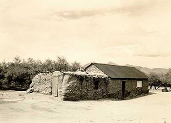 A stone house with a partially thatched roof stands in the dusty yard.  The brush trees can be seen behind the building.