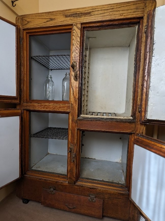 A wooden ice box with all four doors open.  Inside are several racks and glass bottles