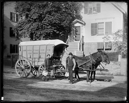 a black and white photo of two men with a horse covered wagon in front of a house