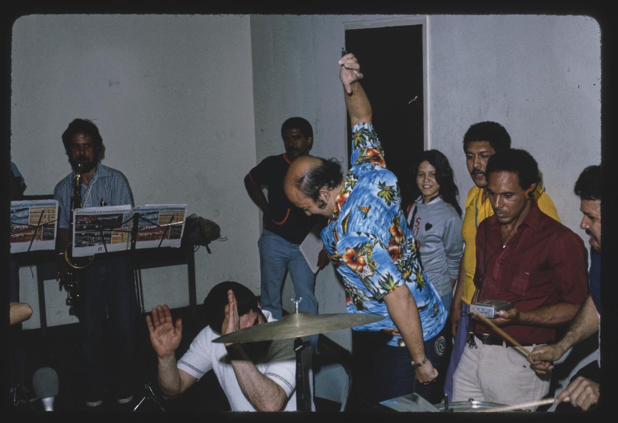 Salsa band in rehearsal with eight people in the frame. Person in the middle is wearing a blue shirt and dancing
