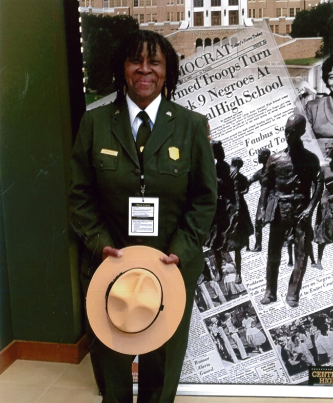 Robin White poses in her NPS uniform in front of an exhibit panel about Little Rock High School. She holds her ranger hat in her hands.