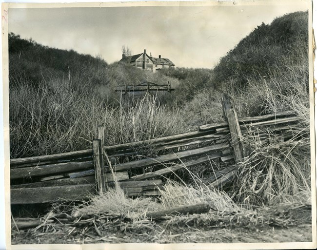 Historic photo of a broken fence with a house at the top of a grassy hill.