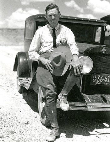 A man wearing breeches sits on the front of his car with his left foot propped up against it.  He holds his hat in his right hand.
