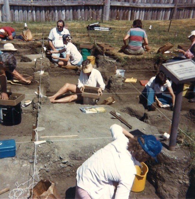 A photo of men and women working in a series of square archaeological units.