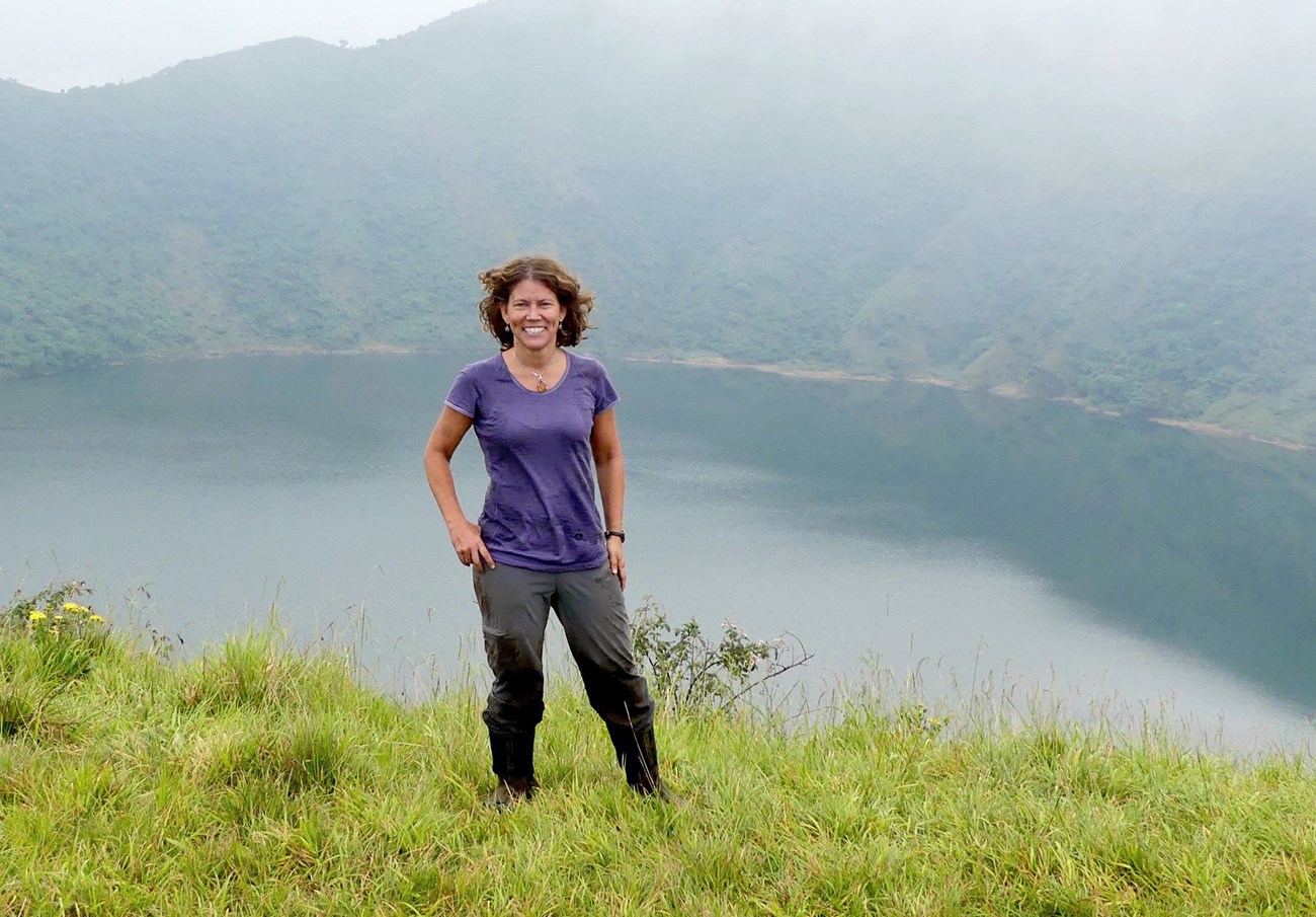 Jena standing in the grass, with peaks and a crater lake behind her.