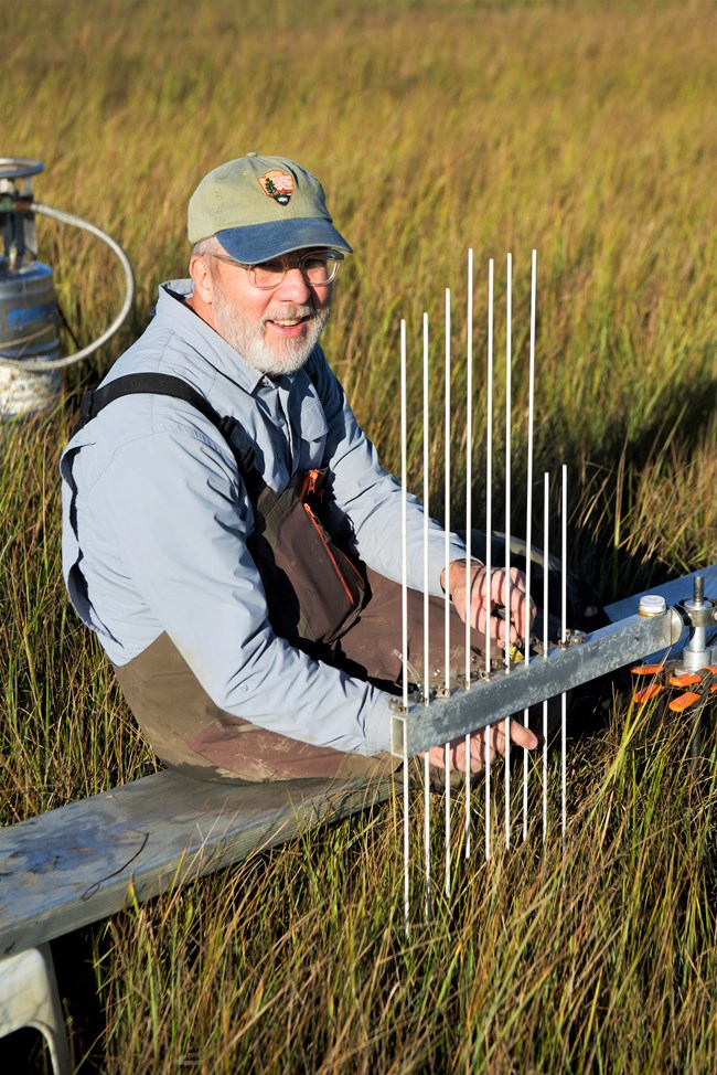 A man in a National Park Service cap kneels in grass to take measurements of a salt marsh at sunset