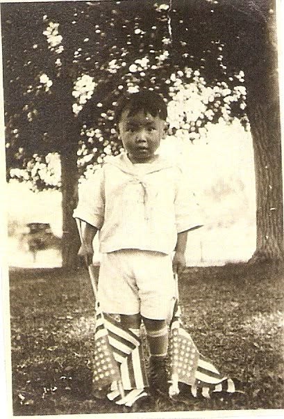 A young child looking straight ahead, holding two small American flags on sticks.