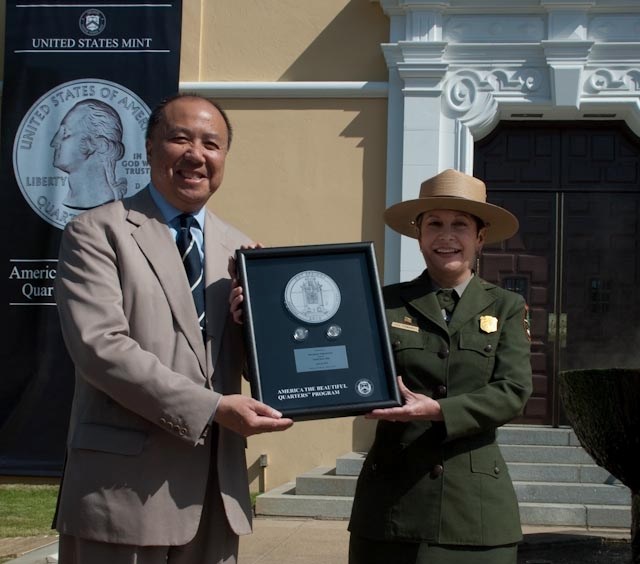 Josie Fernandez in uniform stands with a man in a suit and tie. They each hold onto the sides of a framed NPS quarter set with drawing.