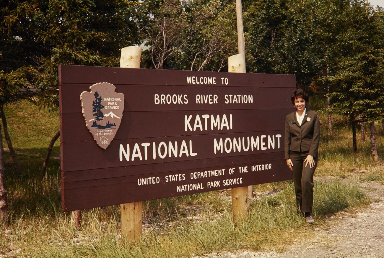 Woman in NPS uniform stands in front of Brooks River sign at Katmai