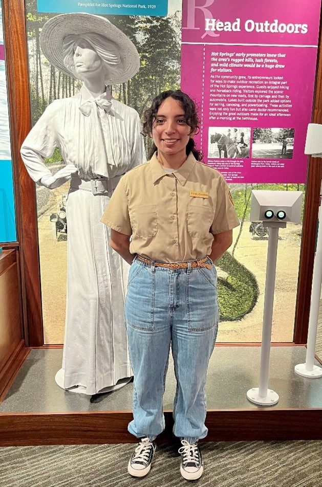 A young woman with dark curly hair wearing a beige button-down shirt and jeans poses with her hands behind her back in front of a white state of a woman in a large hat.