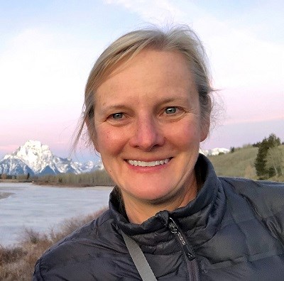 a woman smiles at the camera with snowy peaks in the distance behind her