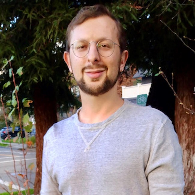 Headshot of a white man with brown hair and a beard wearing glasses and a gray shirt