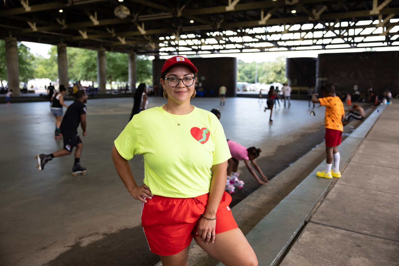 Lydia Daniel smiles for a photo while wearing a light green Friends of Anacostia Park T-shirt on the steps of the rink at Anacostia Park Skating Pavilion in Washington, D.C., on June 28, 2025.