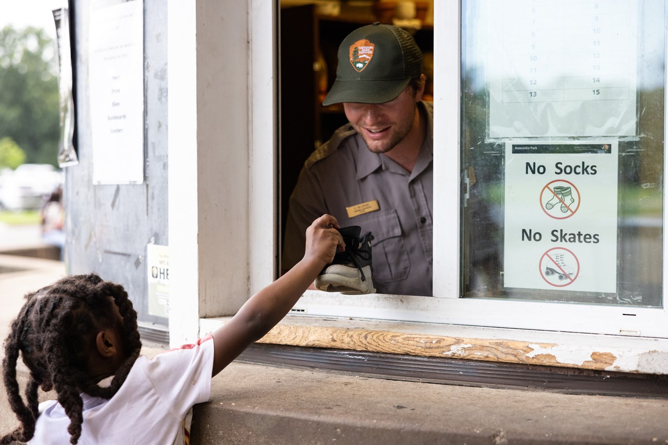 A young boy holds out his sneaker to show his shoe size to smiling National Park Service Ranger Hunter Baldridge at the skate rental booth at Anacostia Park Skating Pavilion in Washington, D.C