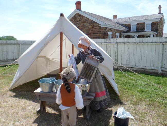 Woman in period clothing demonstrates washing to a child in front of a tent.