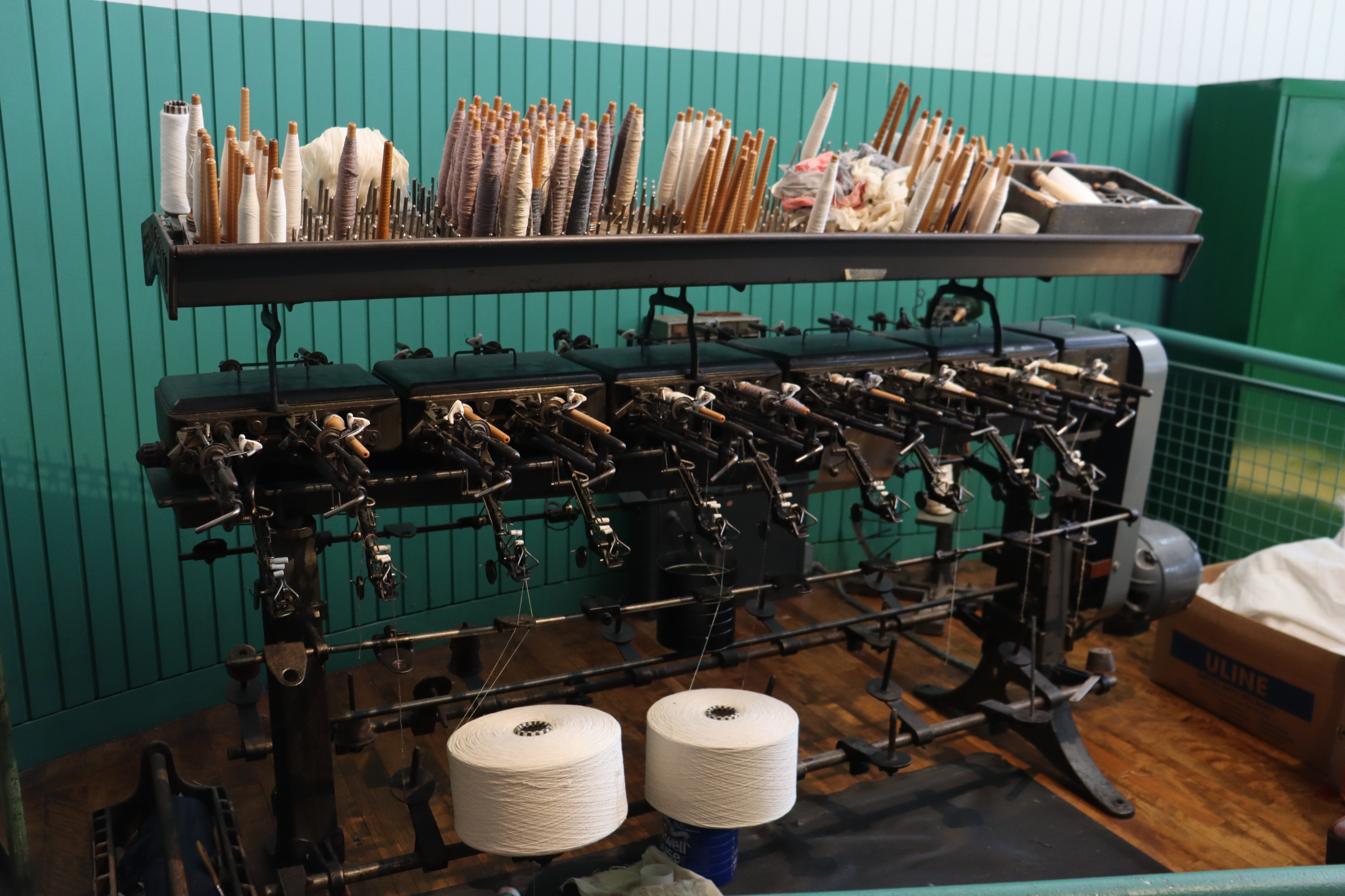 A machine with a rack on the top holding wooden bobbins holding string. Below, bobbins extend out of the machine and string is drawn up from two large rolls of string.