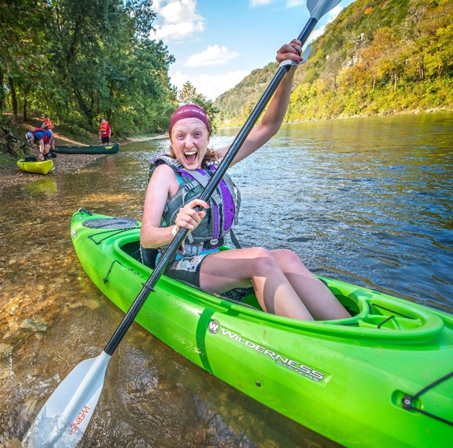 a woman smiles and poses with her kayak oar while sitting in her kayak on the shore of the Buffalo River