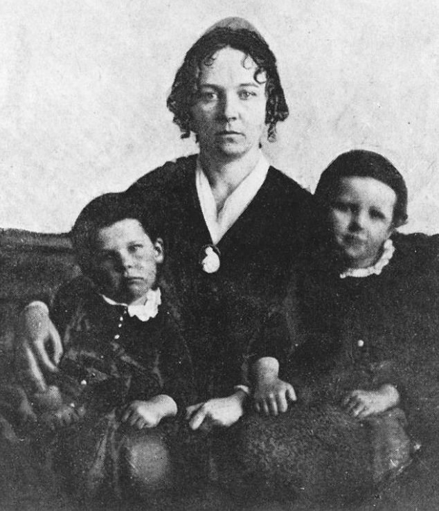 A middle-aged white woman sitting with a young boy on either side of her. They are all dressed in old fashioned clothes.