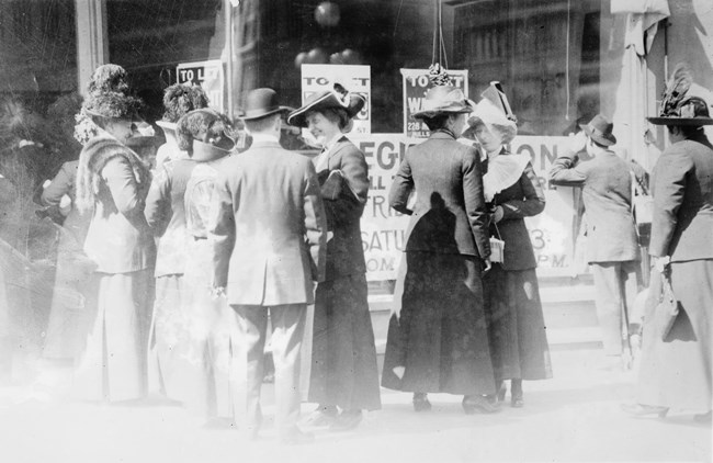 Men and women dressed in 19th century clothing talk to each other. Protest signs are in the background.